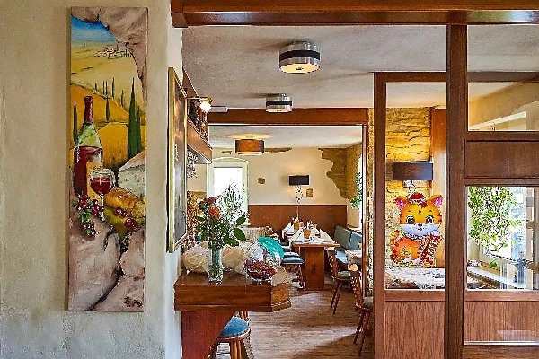Gemütlicher Restaurantbereich mit Holzmöbeln, Wandbild mit Weinflasche links, dekorierter Theke im Vordergrund und heller Sitzecke mit Pflanzen und bunter Winkekatze am Fenster im Hintergrund.