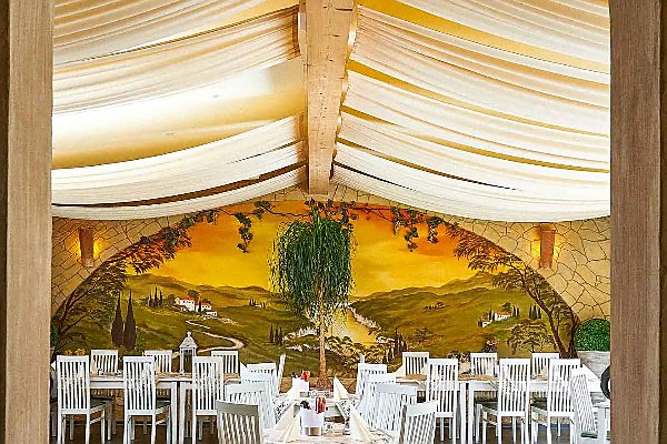 Blick durch einen Rundbogen in einen hellen Restaurantraum mit weiß gedeckten Tischen, weißen Stühlen, heller Stoffdecke und einer großen Wandmalerei einer mediterranen Landschaft im Hintergrund.